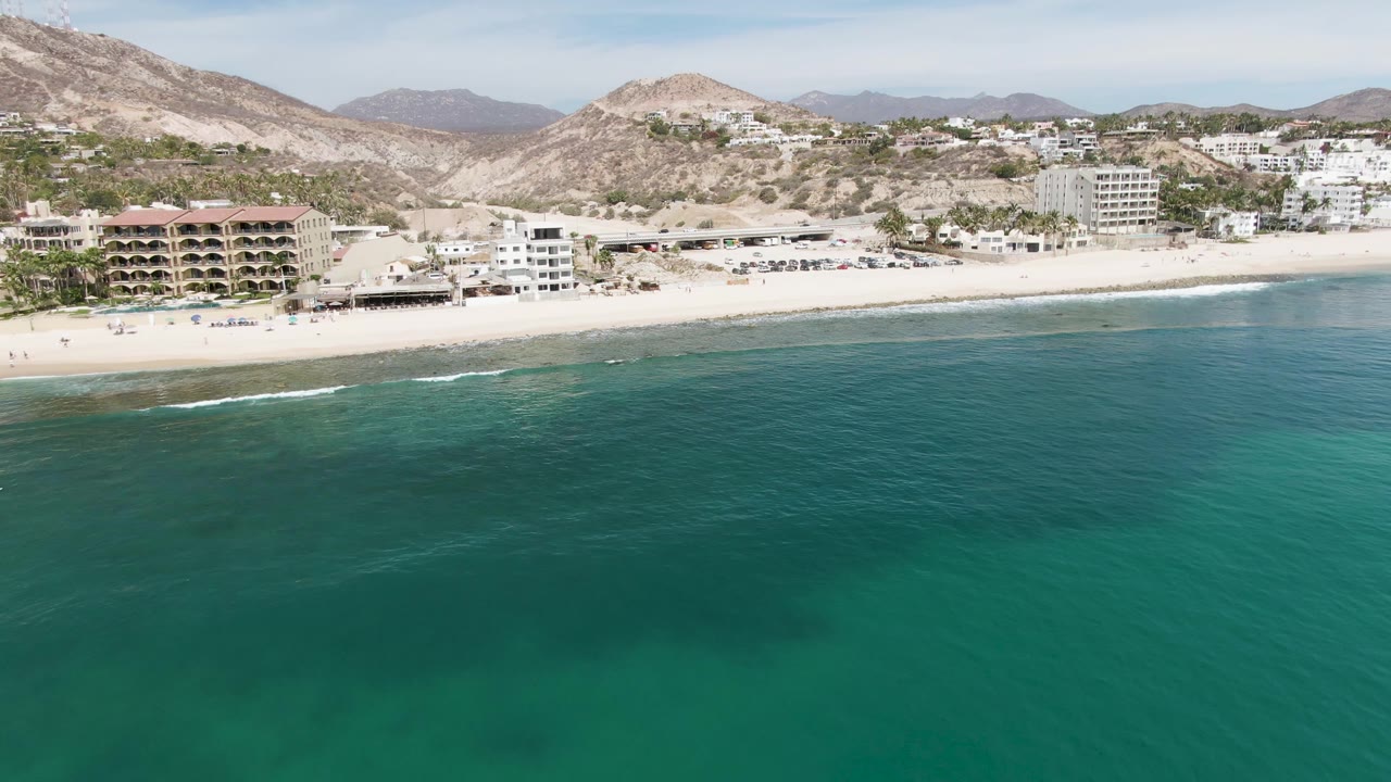 Aerial frame of Punta Gorda Beach with deeper turquoise water in the foreground, a thin surf line, beachfront buildings along the sand, mountain ridges in the background, and no clearly visible people.