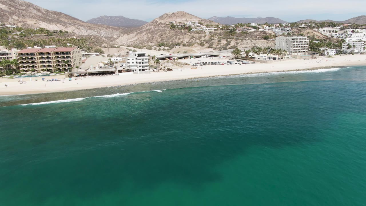 Aerial beach shoreline with green-blue water, beachfront structures, and arid mountains.