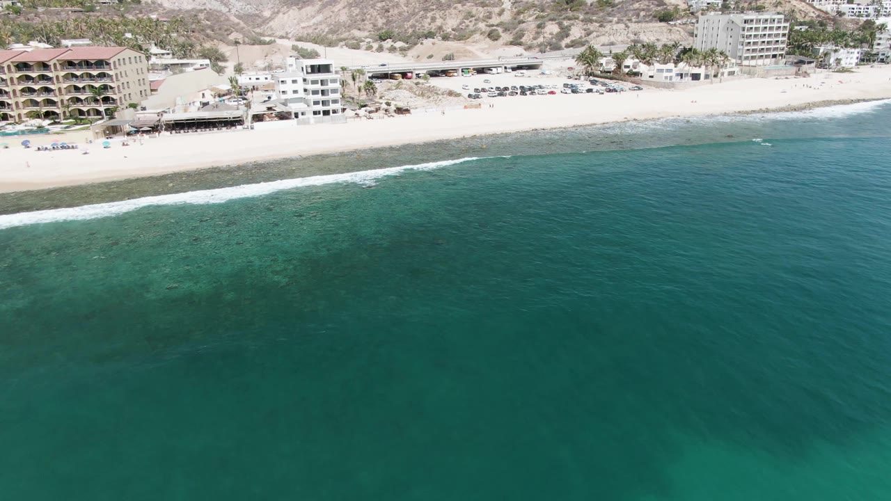 Aerial frame at Punta Gorda Beach, Baja California Sur, showing green-blue ocean in the foreground, a pale beach, coastal buildings, dry hillside terrain, and no visible people.