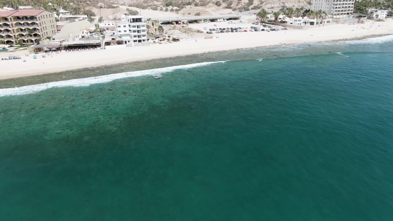 Aerial view over Punta Gorda Beach with darker open water in front, lighter shallow patches near the shore, a narrow surf line, beachfront structures, and no visible people.
