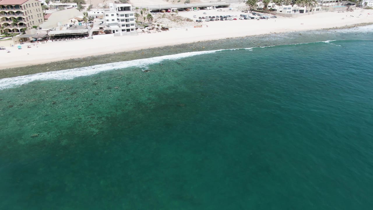 Punta Gorda Beach appears from above with broad teal water, a thin band of white waves, pale sand, multi-story beachfront buildings, parked vehicles, and no visible people.
