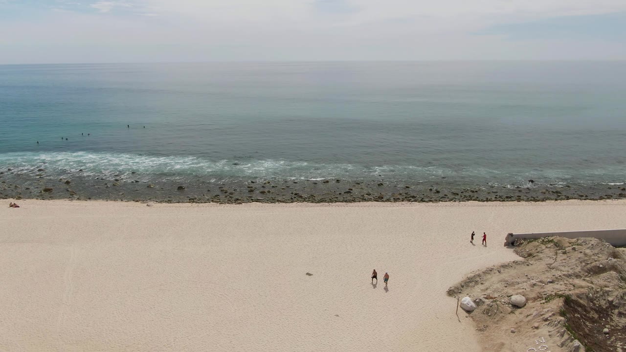 Wide aerial view of Punta Gorda Beach in Baja California Sur, Mexico, with pale sand in front, a rocky surf line, low ocean waves, and small people scattered near the shoreline.