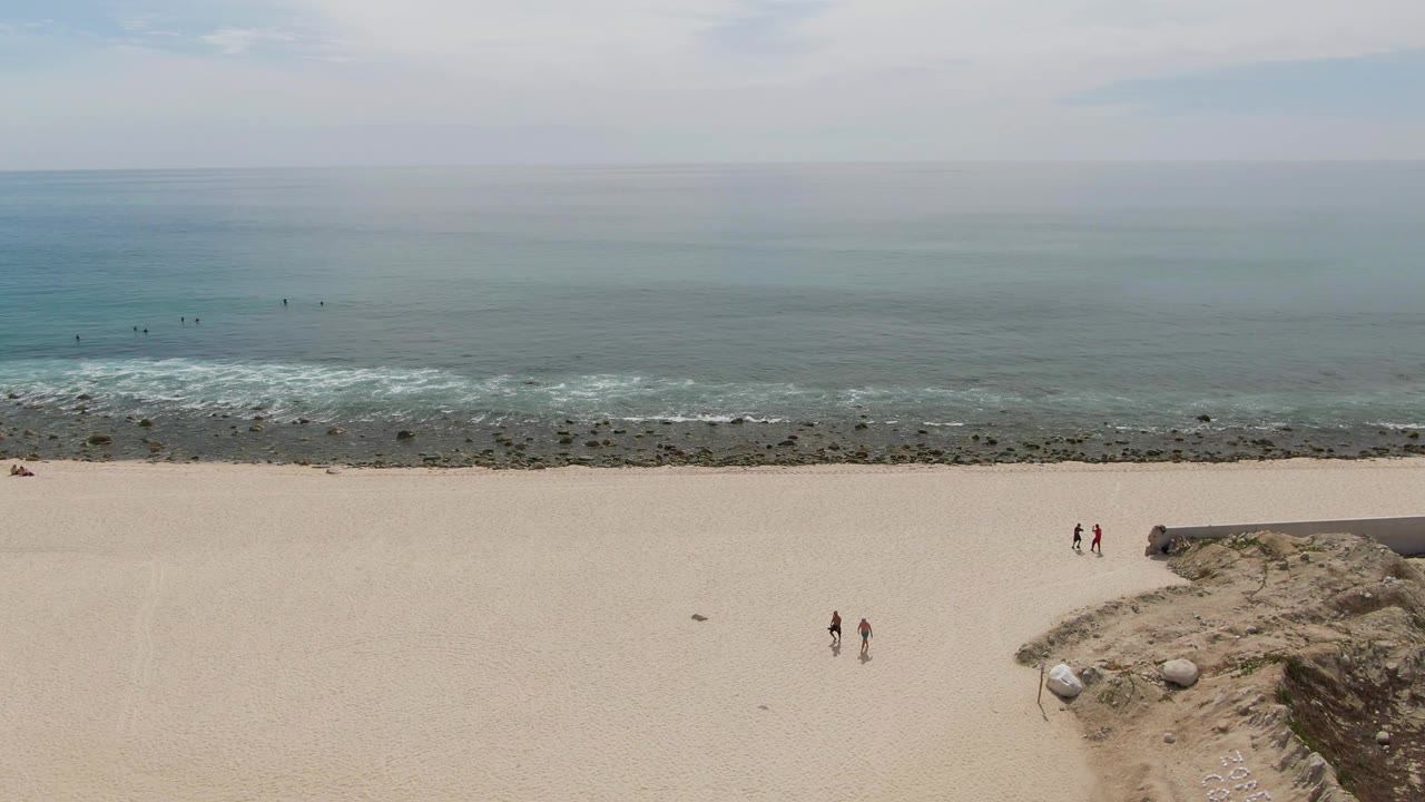 Sandy beach with two people walking, ocean waves, rocks, and hazy sky.