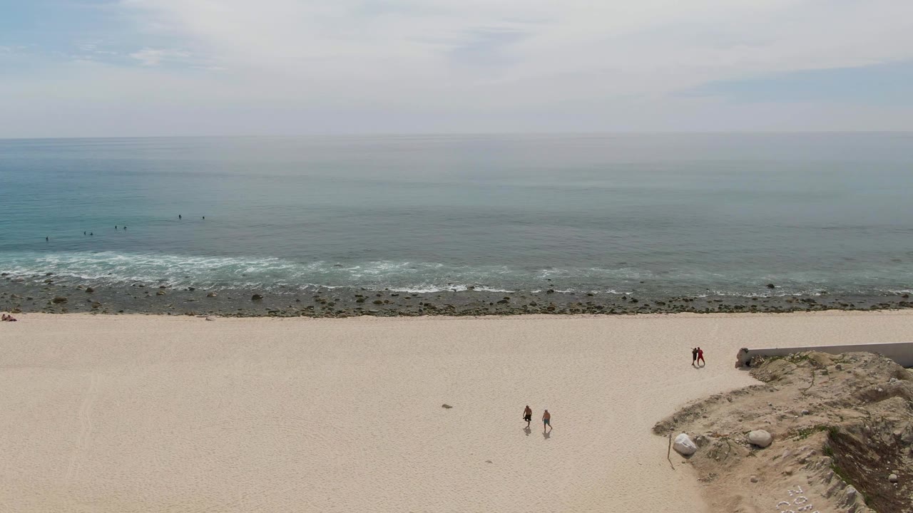 Punta Gorda Beach aerial view with people walking on the sand near a low sandy rise, the rocky shoreline running horizontally, and small white waves breaking across the ocean edge.