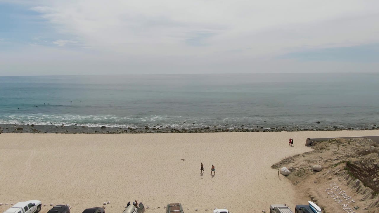 At Punta Gorda Beach in Baja California Sur, a wide elevated frame shows parked vehicles along the sand, two walkers near the center-left, rocky shoreline, low surf, and a hazy partly cloudy sky.
