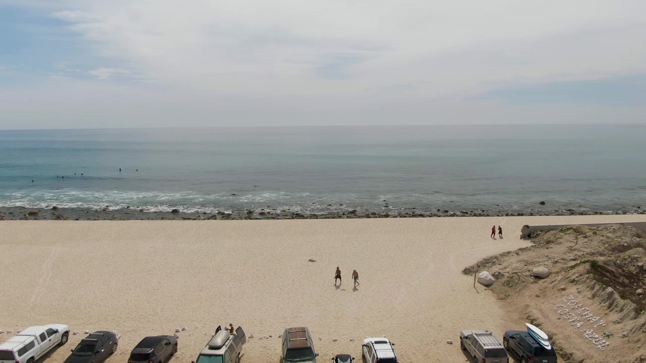 Sandy beach with ocean horizon, parked cars, dunes, and small people walking near the shore.