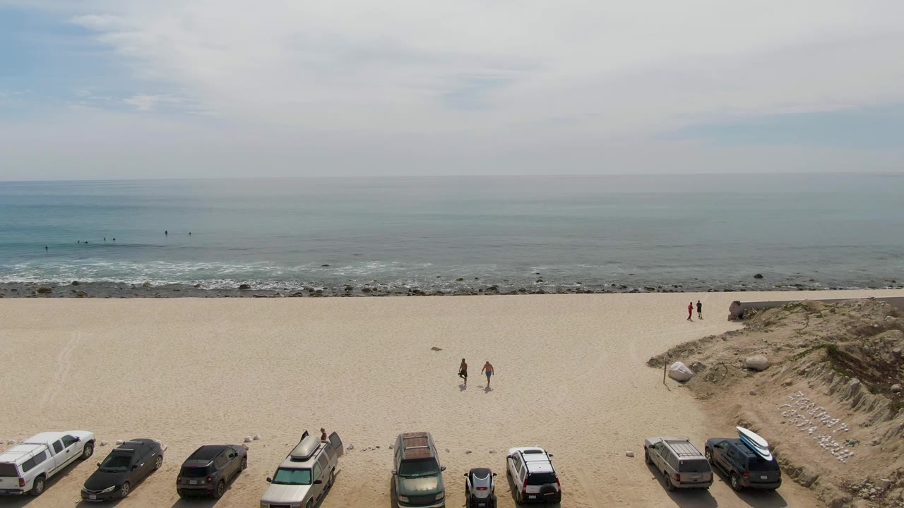 At Punta Gorda Beach in Baja California Sur, parked vehicles line the lower frame beside a sandy dune cut, while walkers move across the beach and small figures remain near the surf.