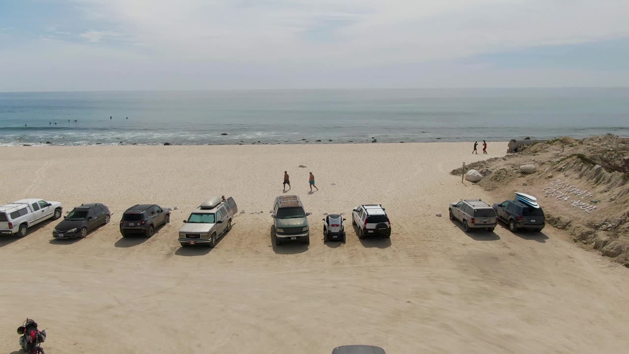 Wide aerial frame over Punta Gorda Beach in Baja California Sur, with parked vehicles lined along the sand, low dunes at both sides, ocean waves in the background, and small people visible near the shore.
