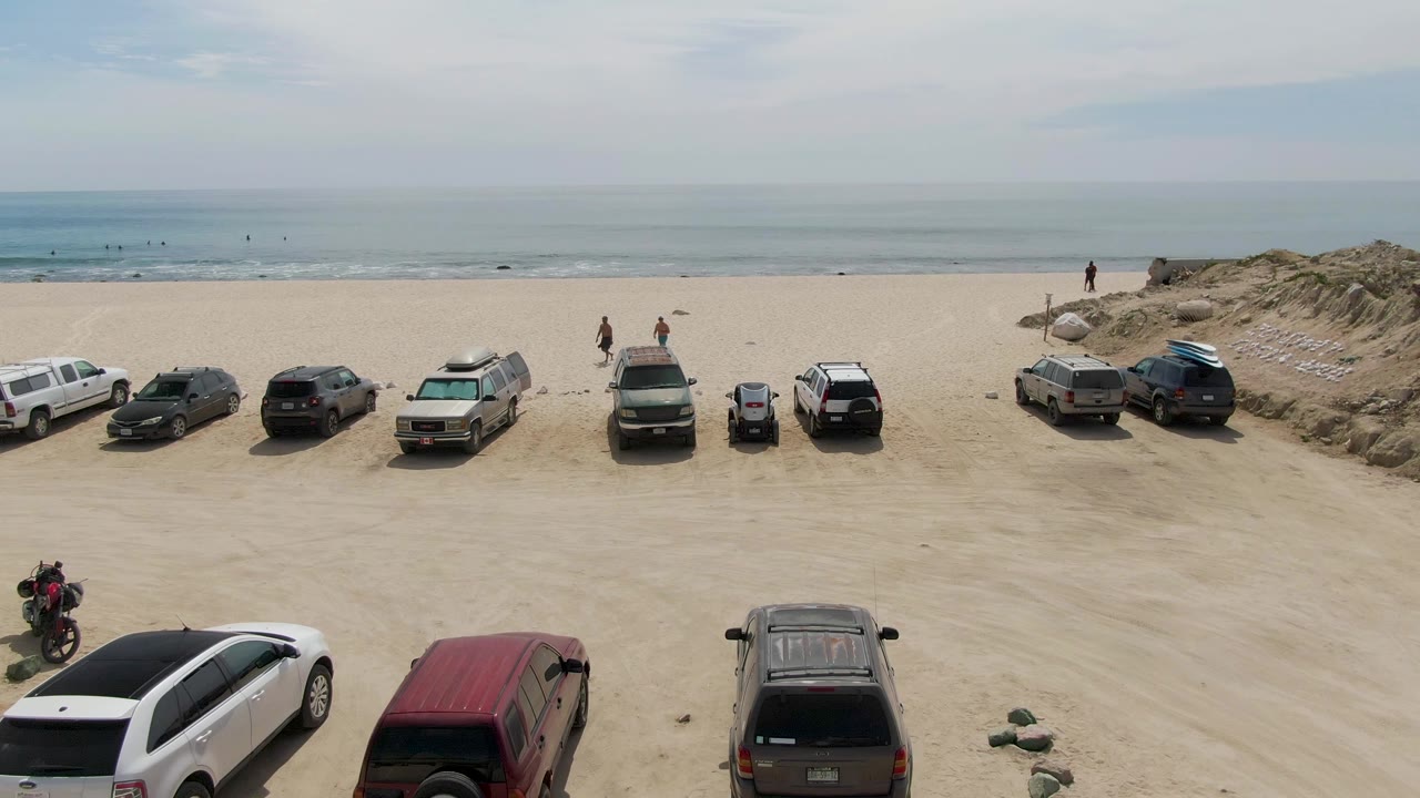 Elevated frame at Punta Gorda Beach showing parked cars spaced across the sand, a low dune ridge behind part of the lot, small figures near the shoreline, and muted blue ocean in the background.