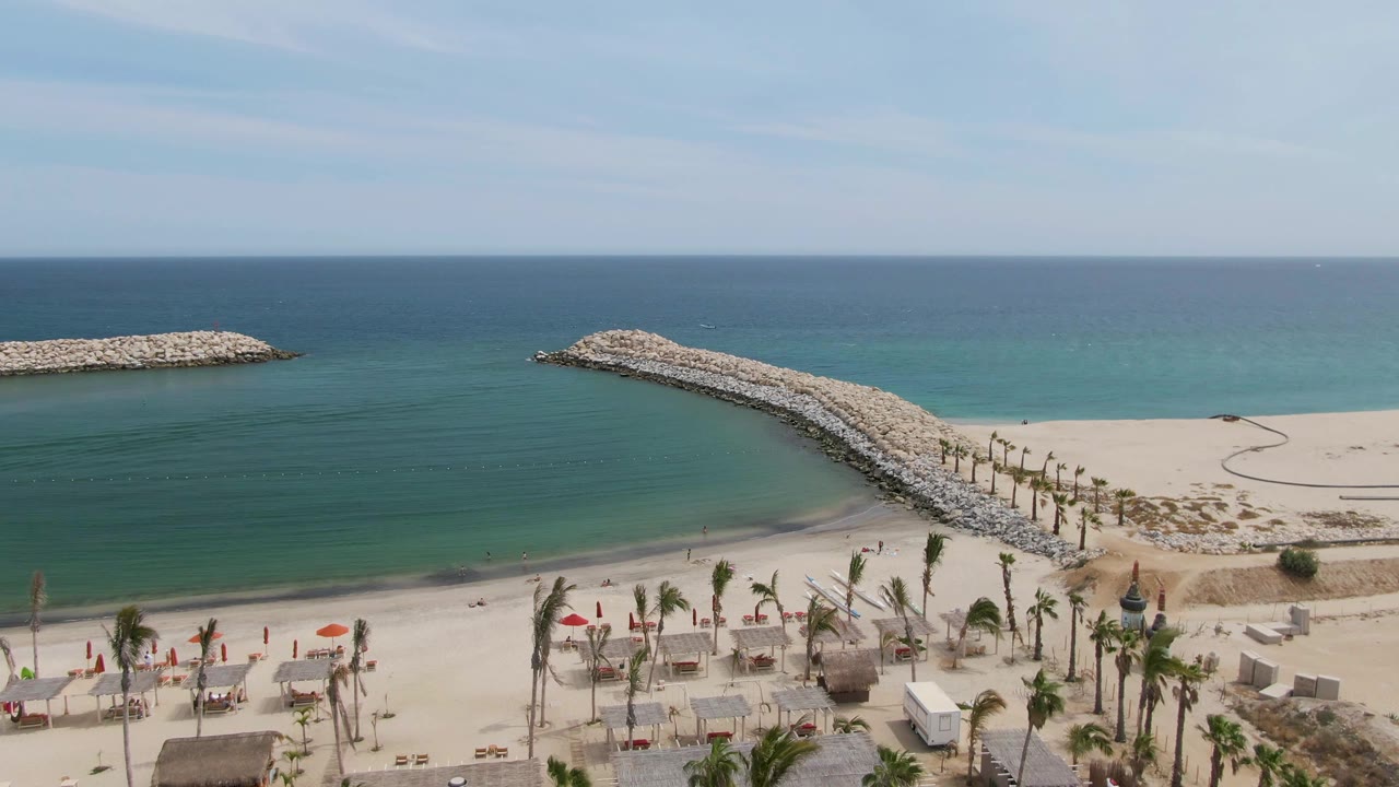 Aerial view over Puerto Los Cabos Beach showing turquoise water, pale sand, a rocky breakwater angled into the sea, beach umbrellas, palms, and no visible people.