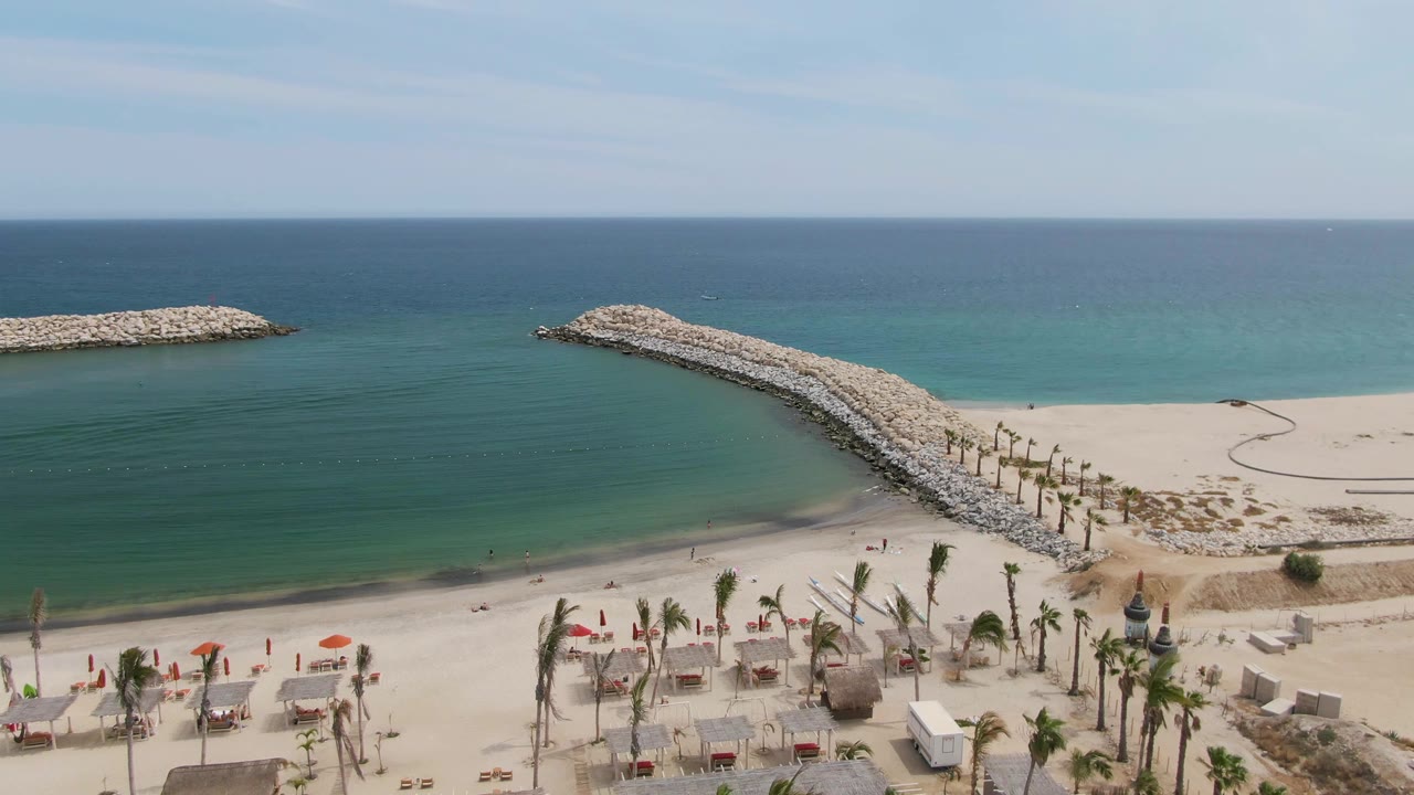 Puerto Los Cabos Beach appears from above with the breakwater centered more prominently, calm green-blue water to the left, open sand to the right, palms below, and no visible people.