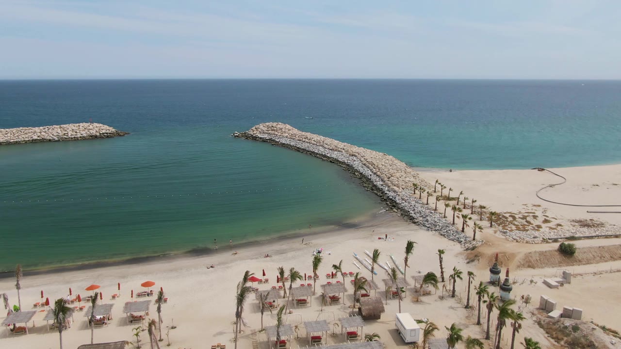The aerial frame shows Puerto Los Cabos Beach with a long rocky breakwater running from the lower right toward the ocean, beach umbrellas and palms along the lower edge, and no visible people.