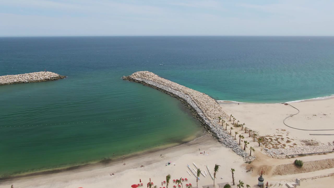 Aerial frame over Puerto Los Cabos Beach showing red umbrellas at the lower left, pale sand, two rocky breakwaters, turquoise shallows, deep blue ocean, and tiny beachgoers.