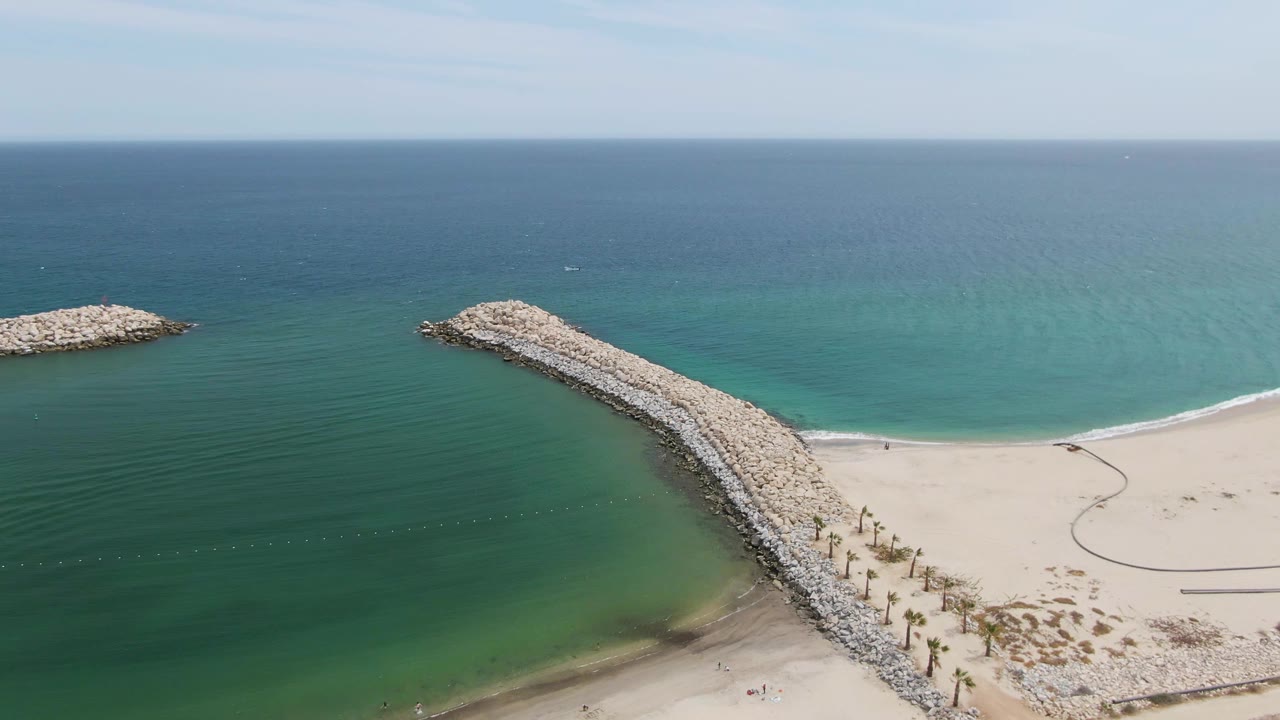 Aerial frame over Puerto Los Cabos Beach showing the curved rock breakwater crossing the center, open sand on the right, green-blue water on the left, ocean horizon above, and a few tiny beachgoers near shore.