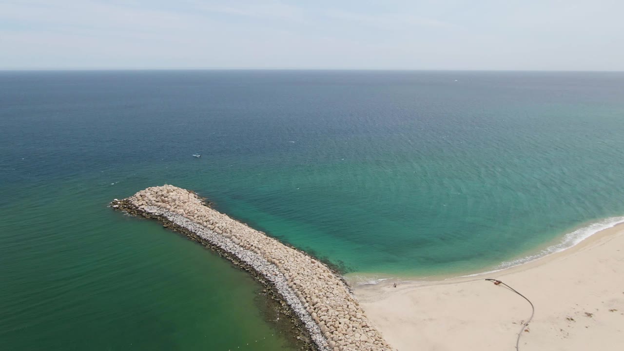 Aerial view at Puerto Los Cabos Beach showing a rock breakwater entering green-blue water beside pale sand, with a high ocean horizon and no visible people.