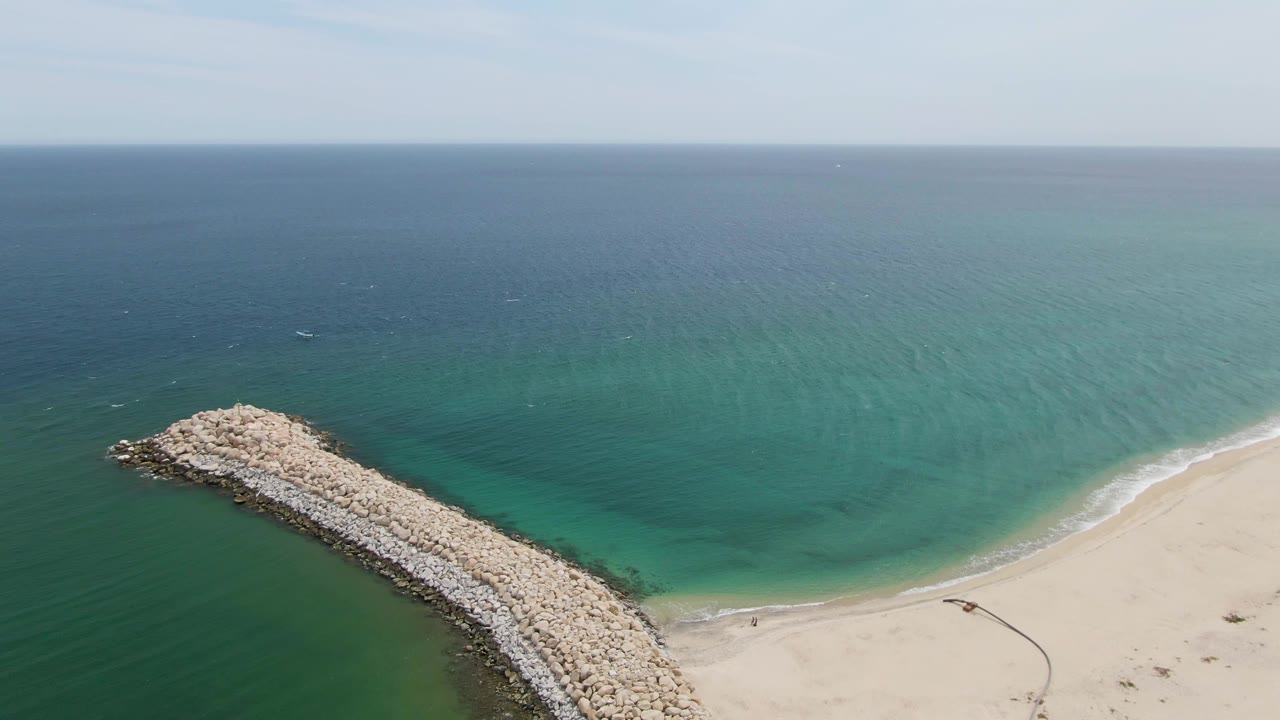 Rock breakwater curves into green-blue water with sandy shoreline on the right.