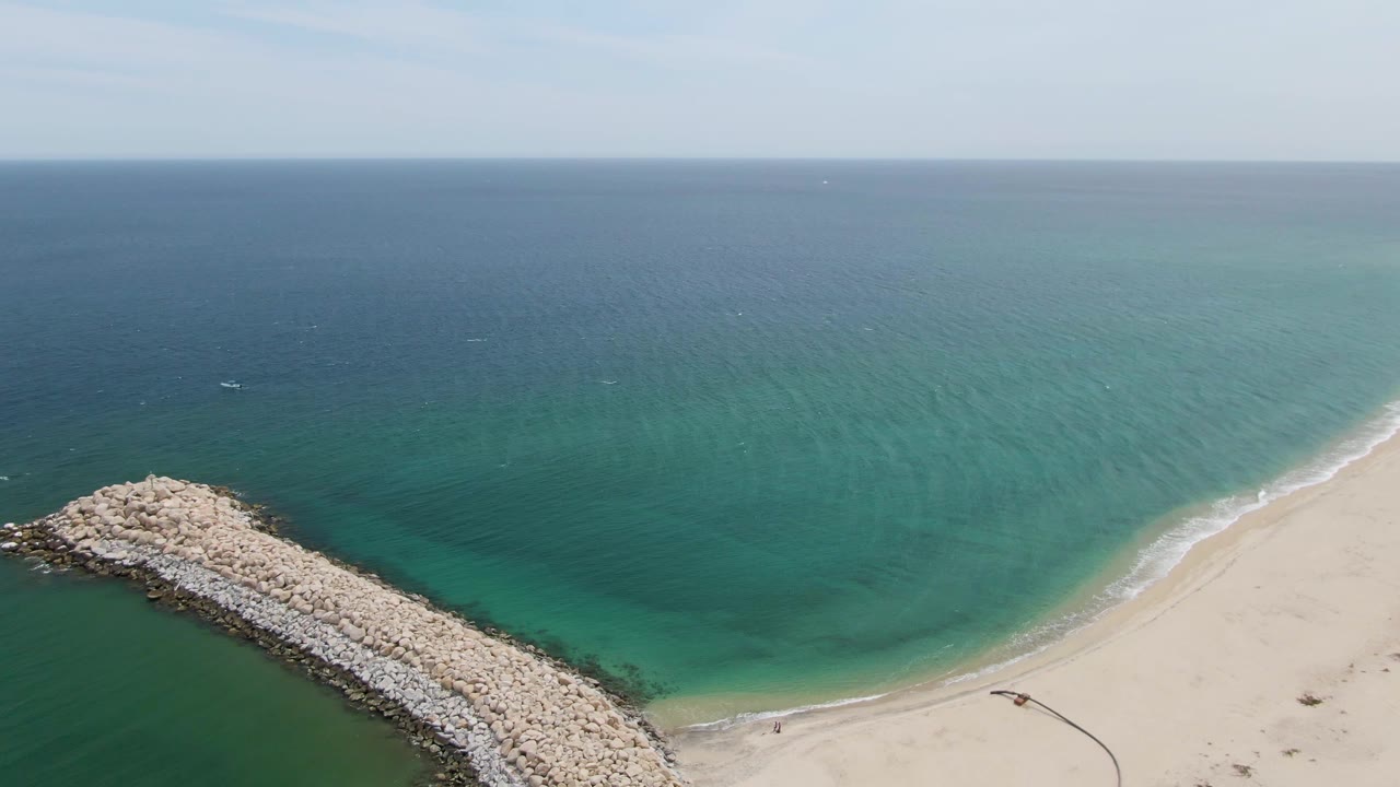 Aerial frame over Puerto Los Cabos Beach with the breakwater near the lower left, a smooth sandy shoreline at right, broad turquoise ocean, and no visible people.