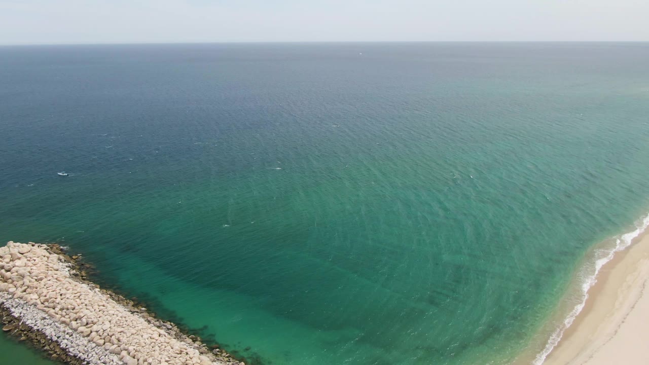 Aerial frame over Puerto Los Cabos Beach showing a pale rock breakwater entering from the lower left, clear green-turquoise water, sandy shoreline, distant horizon, tiny offshore boats, and no visible people.