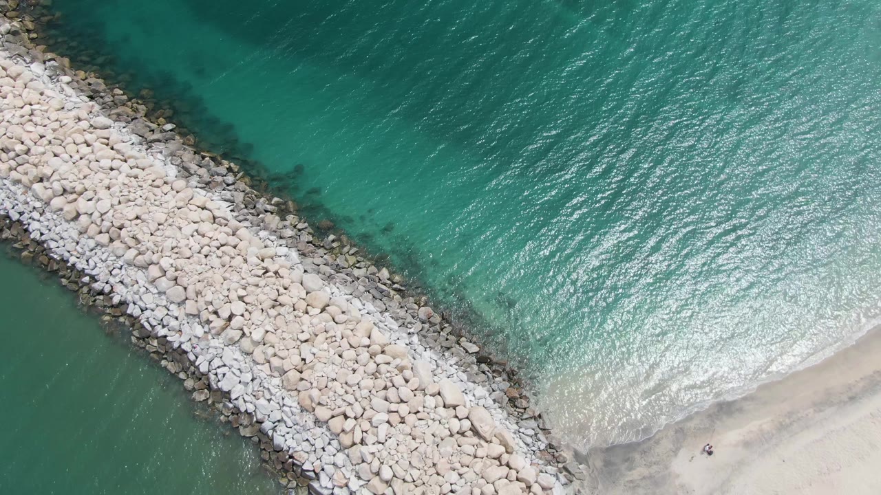 Overhead frame of a pale rock breakwater cutting diagonally through turquoise water at Puerto Los Cabos Beach, with sand visible at the lower right and no visible people.