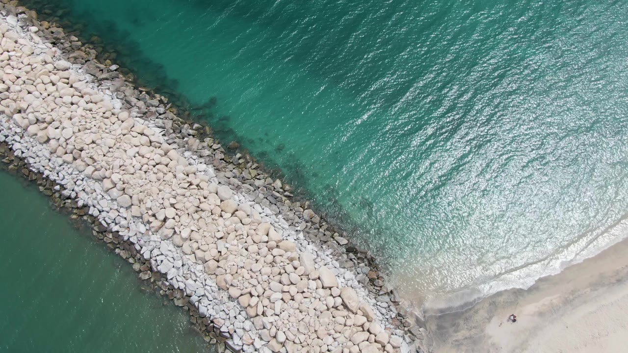 Puerto Los Cabos Beach aerial frame showing pale breakwater stones, bright turquoise surface texture, and a small white wave advancing onto the sandy shore with no visible people.