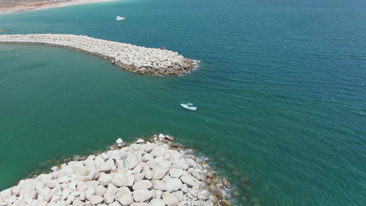 Aerial view at Puerto Los Cabos Beach with the foreground rocks filling more of the lower edge, a small boat in the green water, a long breakwater beyond it, and no visible people.