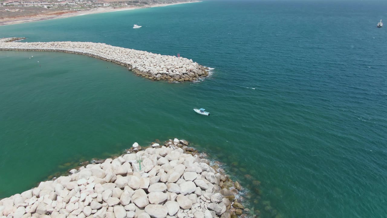 Aerial frame over Puerto Los Cabos Beach showing a rounded foreground breakwater, a small boat on calm blue-green water, a sandy coast in the background, and no visible people.
