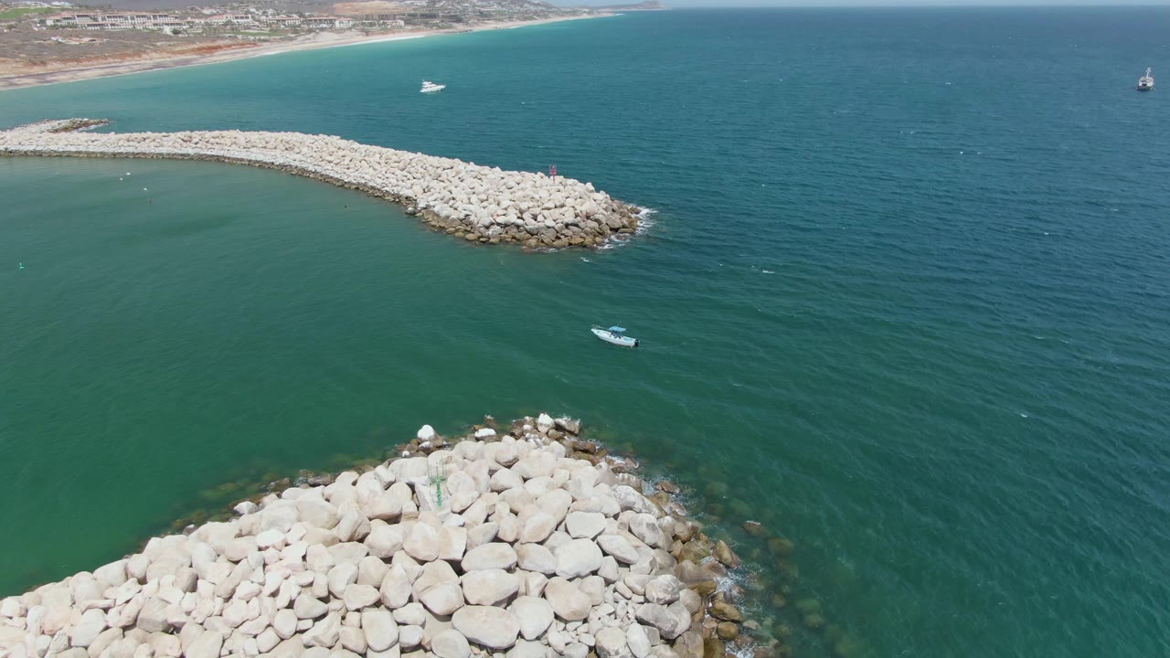 Puerto Los Cabos Beach aerial frame with the foreground rock pile low in frame, the long breakwater stretching across the upper left water, the small boat below it, and no visible people.