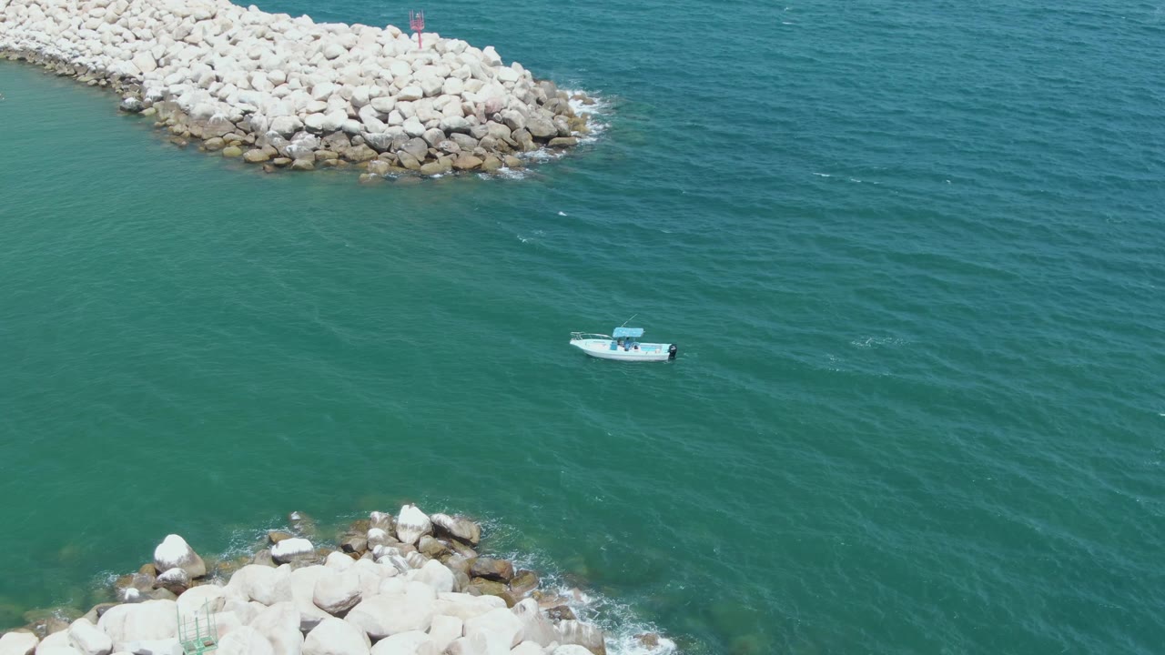 At Puerto Los Cabos Beach, a small white boat sits in the channel between pale rock breakwaters, with rippled green-blue water and no visible people.