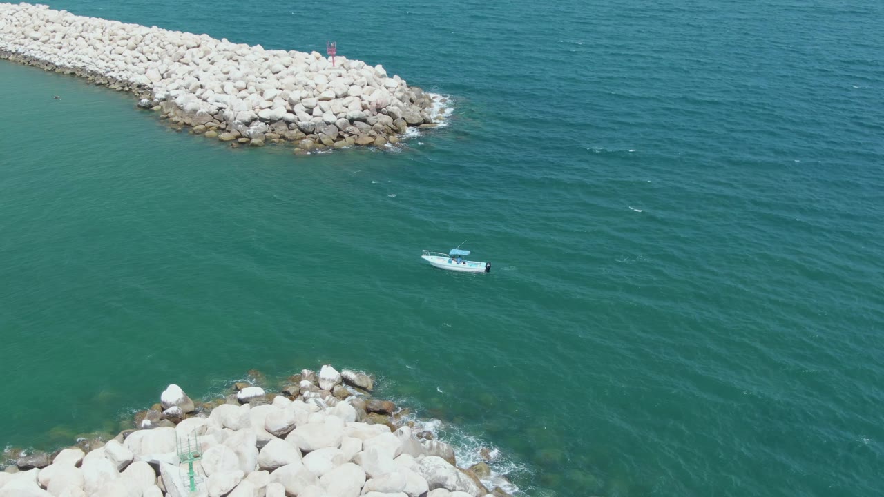 At Puerto Los Cabos Beach, the boat remains below the long pale boulder breakwater, with darker ripples spreading across the open water and no visible people.