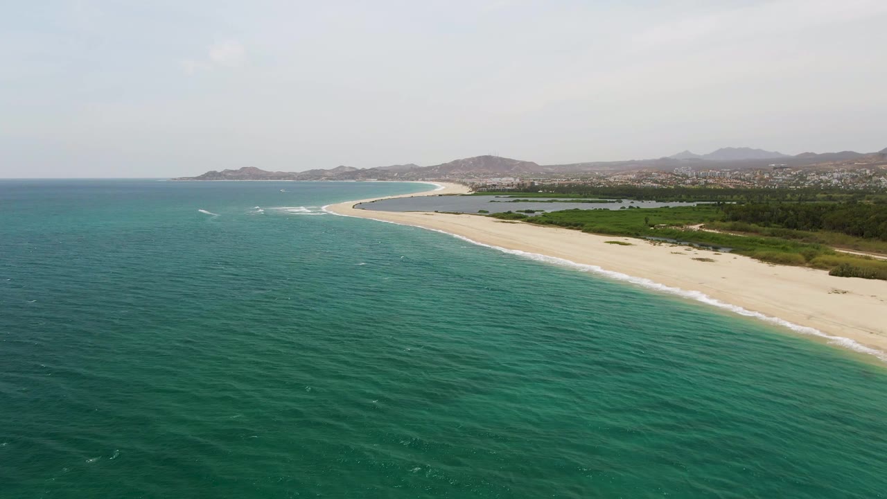 Aerial frame over Puerto Los Cabos Beach showing turquoise water, a pale curving shoreline, low greenery, distant buildings and hills, and no visible people.