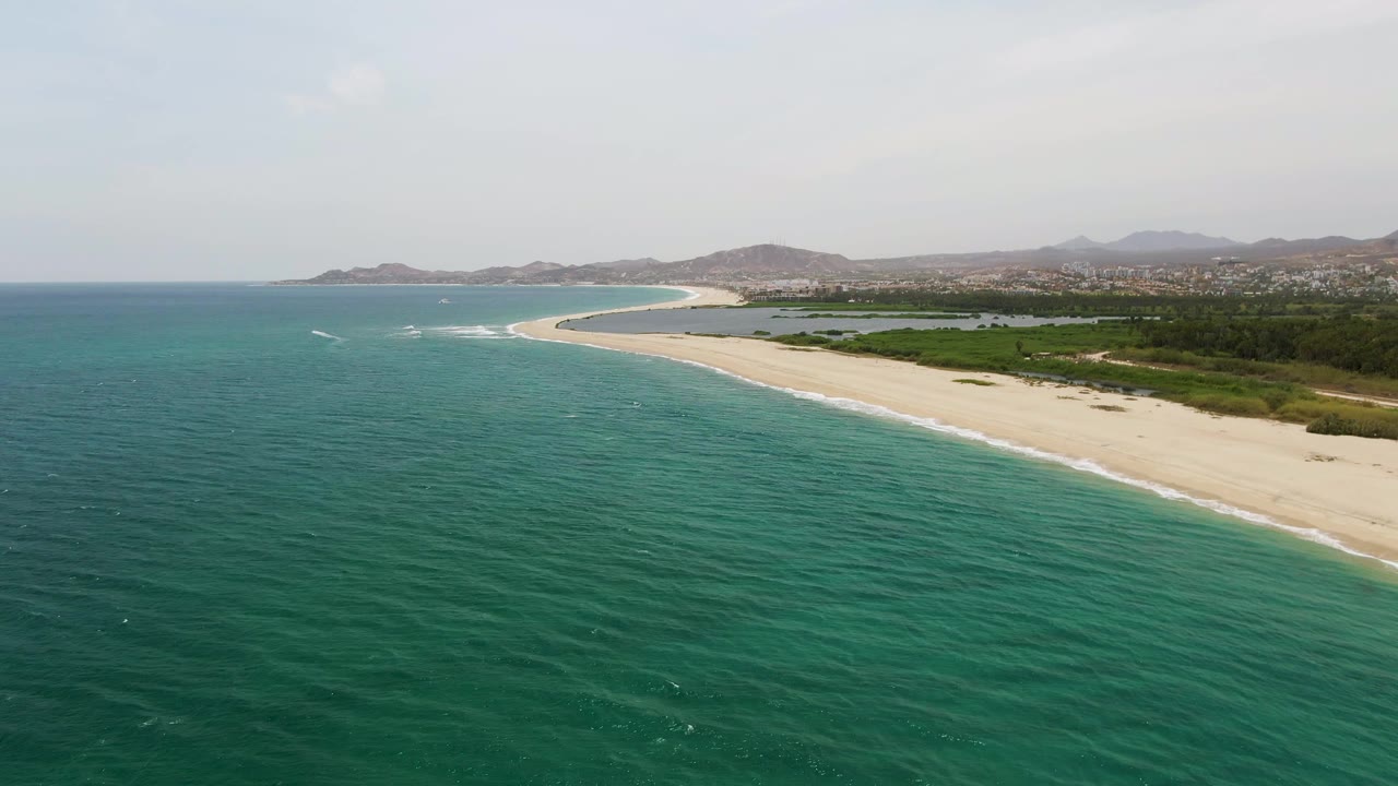 A second aerial view of Puerto Los Cabos Beach keeps the shoreline high in frame, with rippled turquoise water below, pale sand, inland greenery, distant hills, and no visible people.