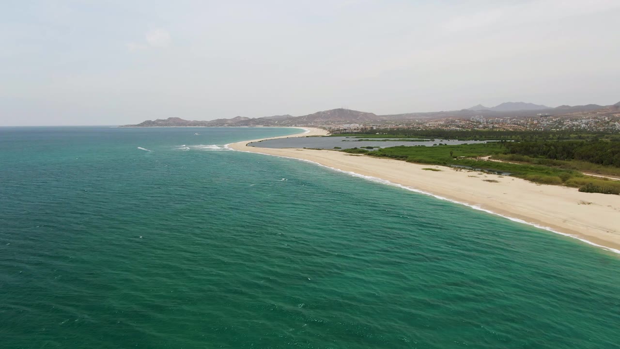 Puerto Los Cabos Beach appears from an elevated angle with more open turquoise water in the foreground, the sand band curving along the right side, low greenery, hills, and no visible people.