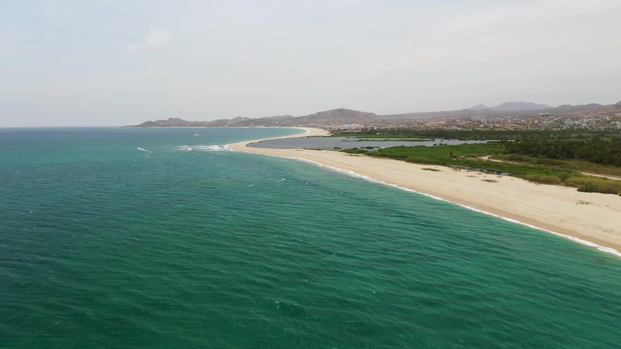 Aerial beach view with white surf lines along sand and hills under a hazy sky.