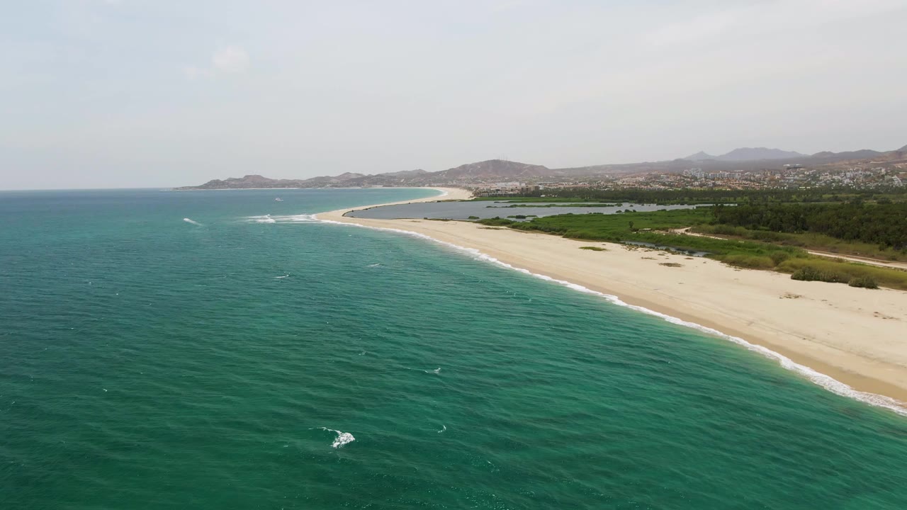 Aerial view of turquoise ocean beside a wide sandy shoreline and distant hills.