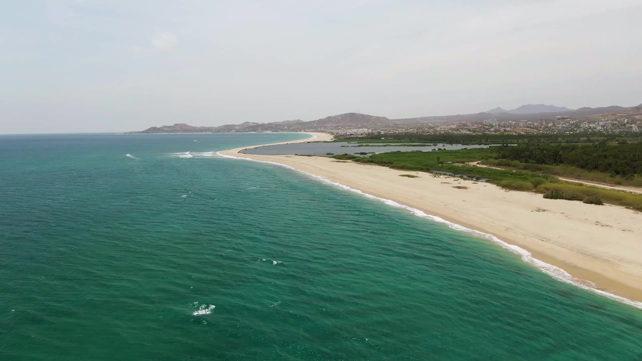 At 0:01, Puerto Los Cabos Beach remains in a wide aerial composition, with whitecaps scattered across the turquoise water, the sandy beach running diagonally, inland greenery, distant hills, and no visible people.