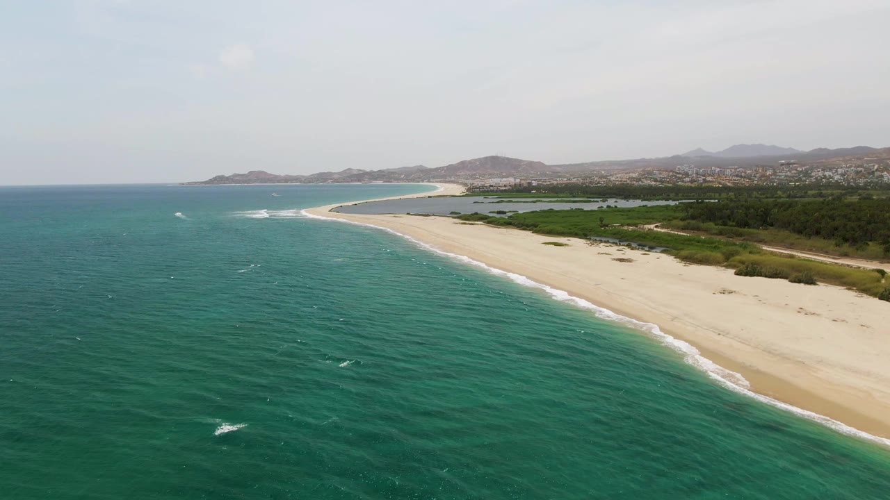 At 0:02, the aerial view of Puerto Los Cabos Beach shows more open water in the foreground, thin surf along the pale sand, a green band behind the beach, low hills on the horizon, and no visible people.