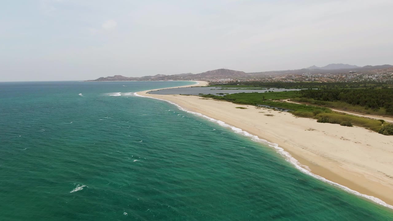 At 0:03, Puerto Los Cabos Beach is seen from above with a longer stretch of surf line visible, turquoise water filling the left foreground, pale sand on the right, inland greenery, distant hills, and no visible people.