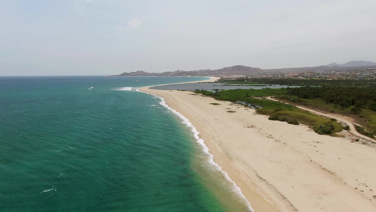 At 0:02, the aerial angle over Puerto Los Cabos Beach shows more open water at left, a narrow band of foam, pale sand, inland greenery, and no visible people.