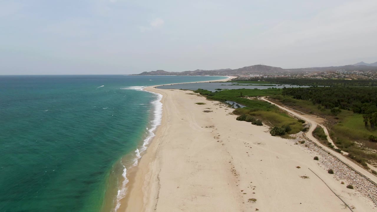 Aerial view over Puerto Los Cabos Beach with turquoise ocean at left, a wide sandy shoreline, a dirt coastal path at right, distant hills, and no visible people.