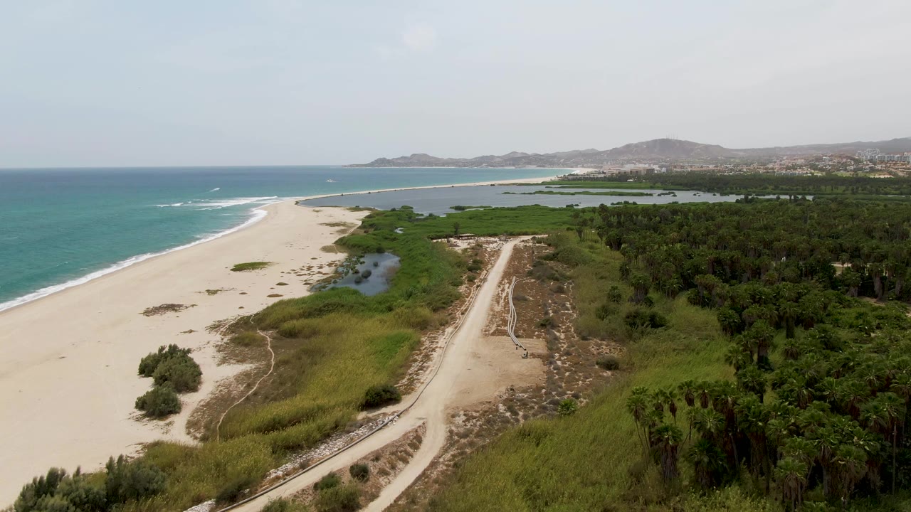 Aerial frame over Puerto Los Cabos Beach, Mexico, showing turquoise ocean beside pale sand, a curving dirt path through dunes, inland greenery, distant hills, and no visible people.