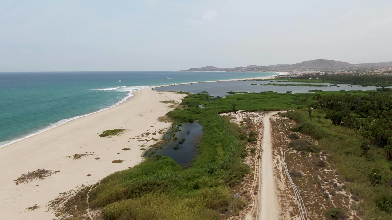 Puerto Los Cabos Beach aerial view with the shoreline curving left, lagoon water and dense greenery in the middle, a pale dirt road on the right, and no visible people.