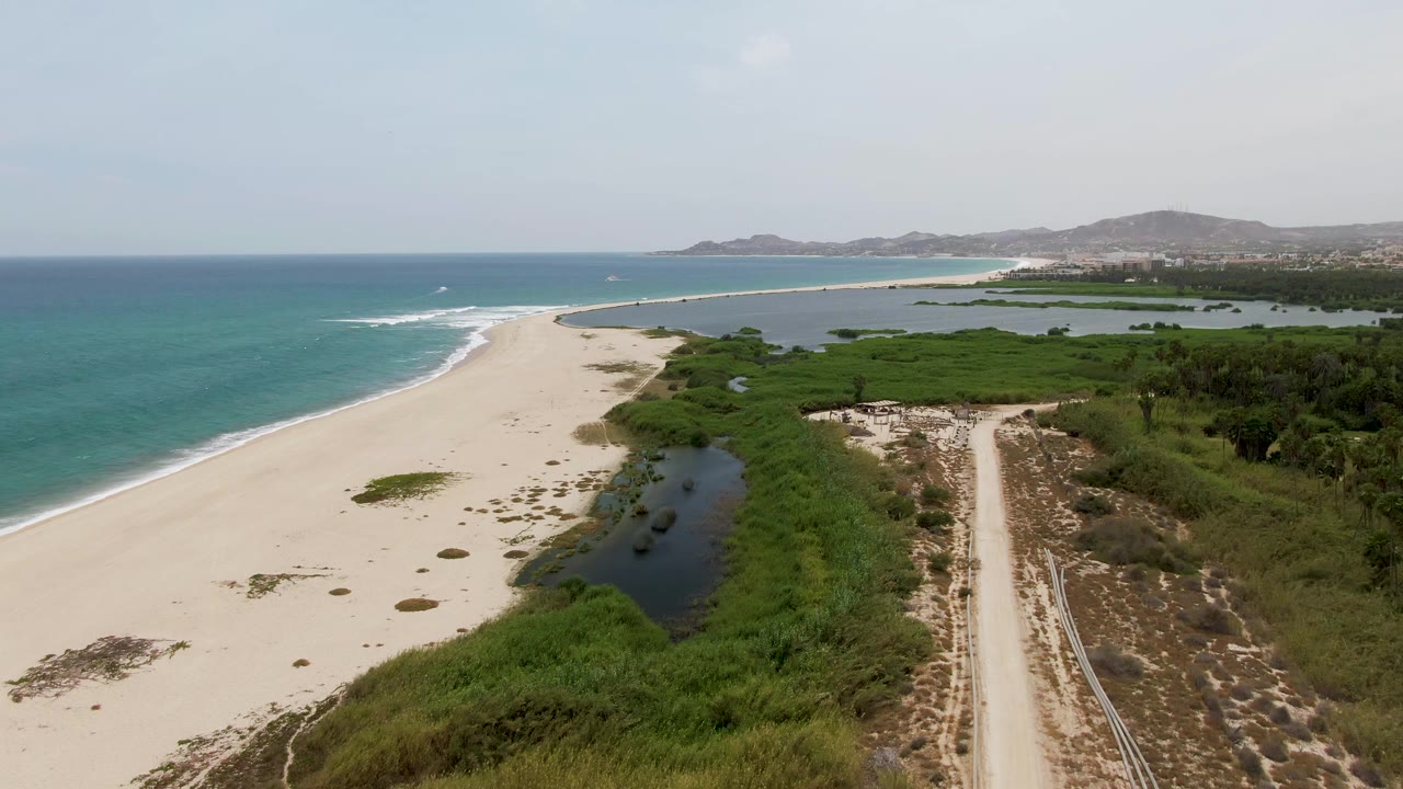 Aerial view at Puerto Los Cabos Beach showing a dark lagoon pool bordered by green plants, turquoise ocean on the left, dirt road on the right, distant hills, and no visible people.