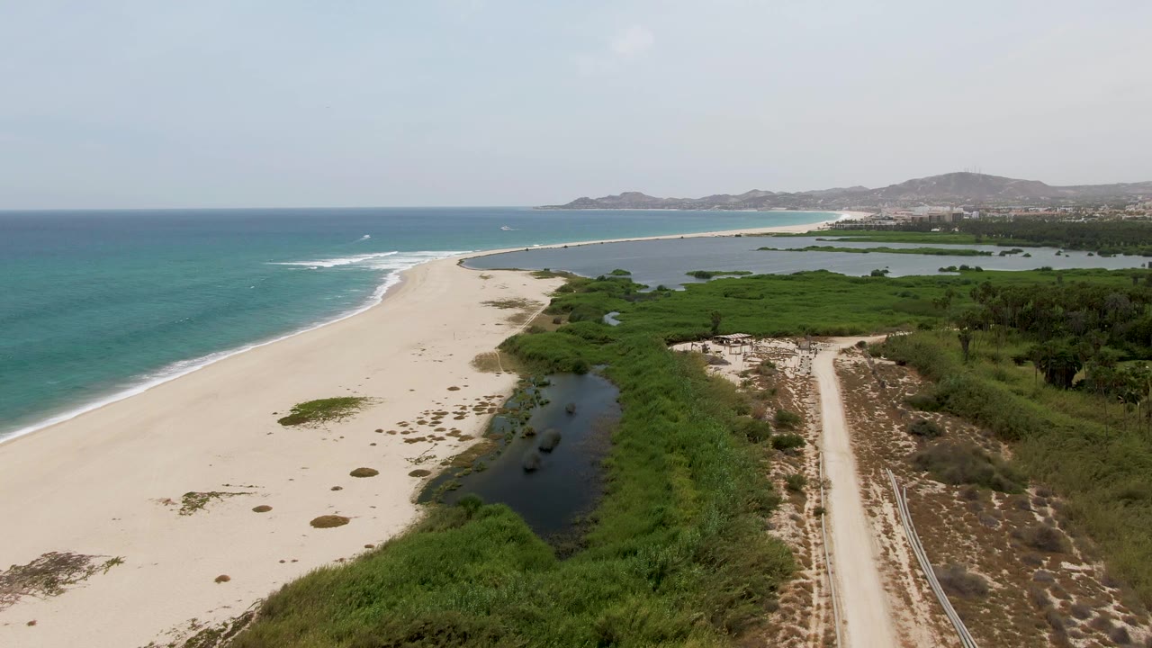 Puerto Los Cabos Beach aerial frame with more open sand visible in the foreground, ocean surf running along the left edge, lagoon vegetation near center, a dirt road at right, and no visible people.