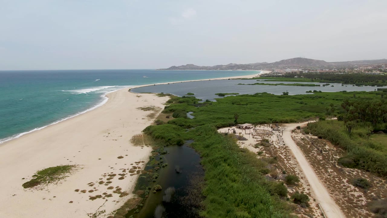 Aerial frame of Puerto Los Cabos Beach with the shoreline curving into the distance, lagoon water beside dense vegetation, a pale dirt track at right, and no visible people.