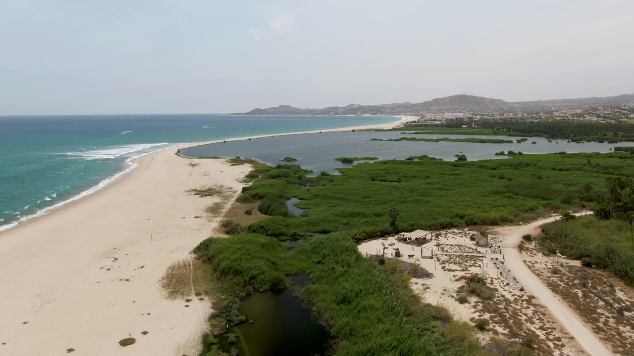 Aerial frame of Puerto Los Cabos Beach with pale sand on the left, turquoise ocean waves, a green lagoon behind the beach, dense coastal vegetation, distant hills, and no visible people.