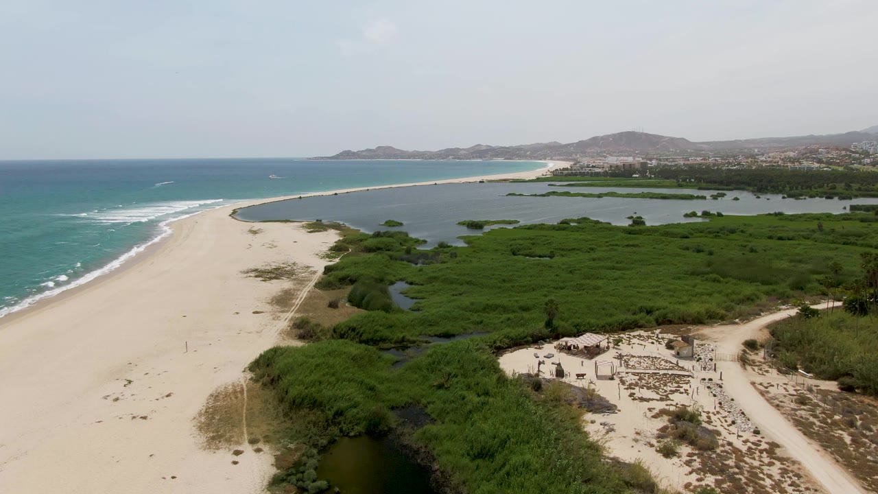 Drone view over beach, lagoon water, marsh greenery, and small structures near shore.