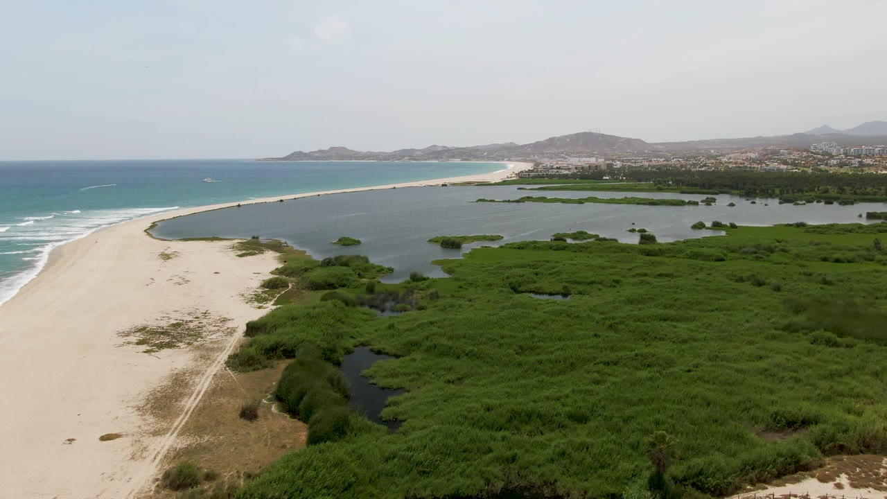 Aerial frame at Puerto Los Cabos Beach shows the sand path slightly farther along the lagoon edge, with ocean surf left, mangrove wetland right, distant shoreline, and no visible people.
