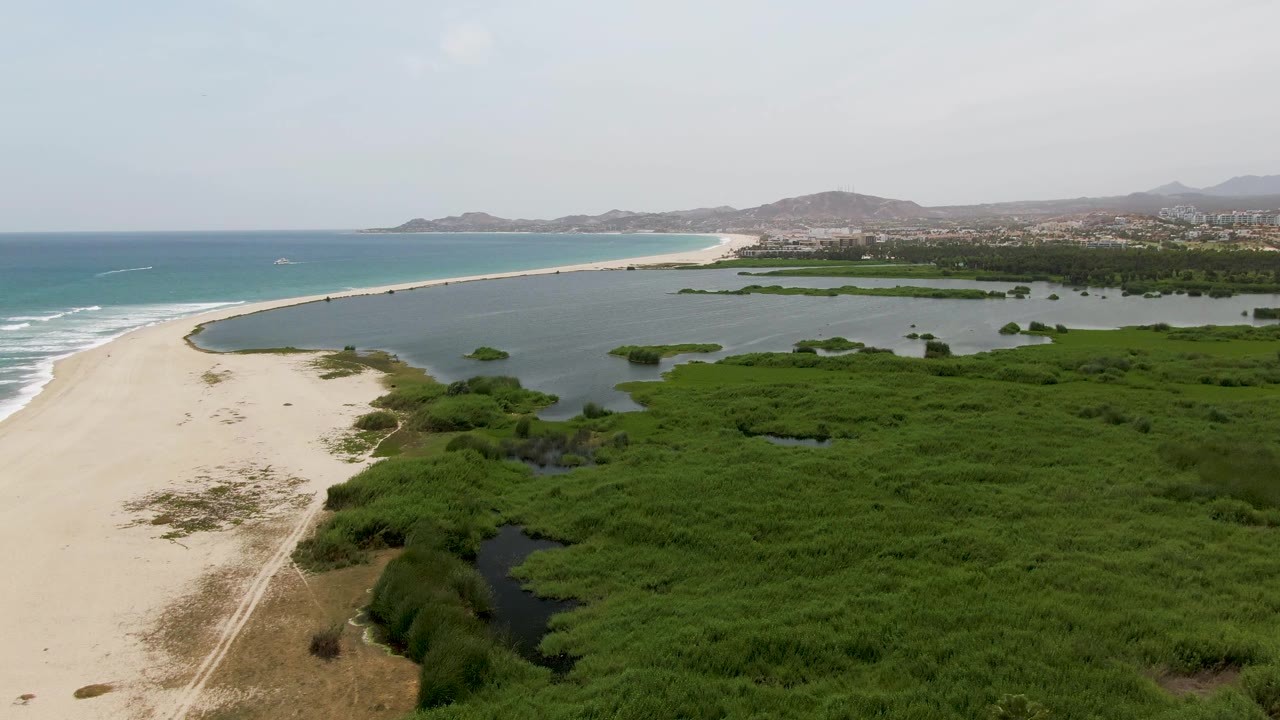 Aerial frame at Puerto Los Cabos Beach keeps the lagoon broad in the midground, with green mangroves filling the lower right, ocean waves along the left edge, and no visible people.