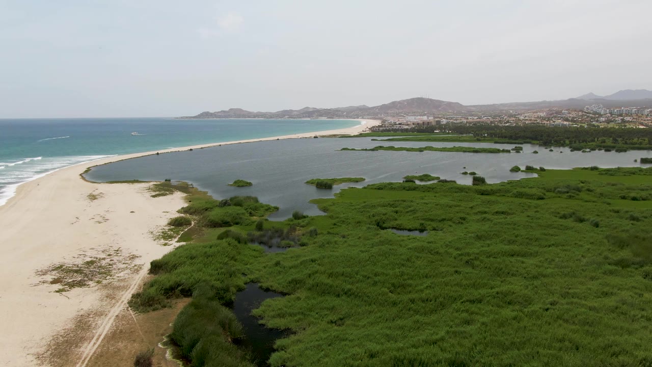 Aerial frame at Puerto Los Cabos Beach looks along the sandy beach with surf to the left, a dark-edged lagoon to the right, green wetland growth below, distant hills, and no visible people.