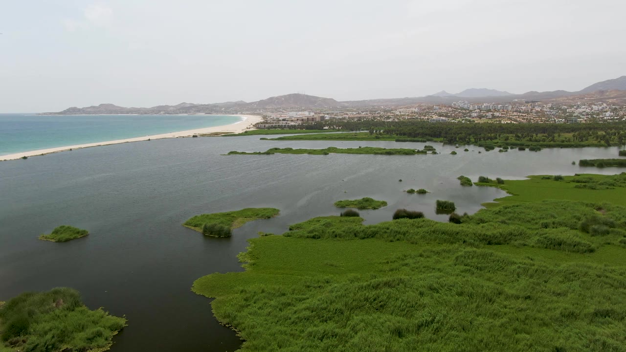 A second Puerto Los Cabos Beach aerial shows marsh islands spread across calm lagoon water, with the sandy beach and blue-green ocean at left, low hills beyond the settlement, and no visible people.